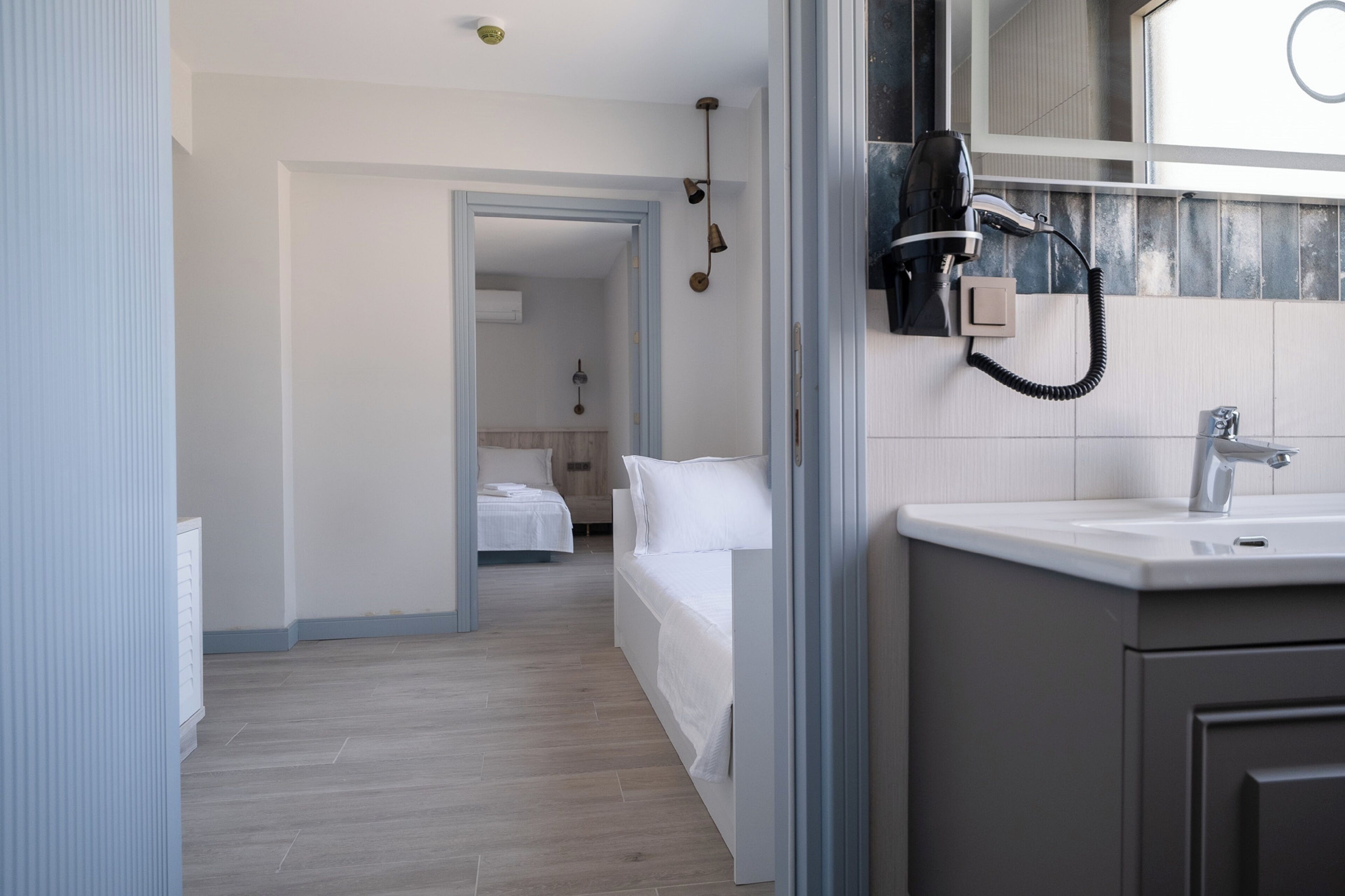 Une chambre d’hôtel moderne avec un grand miroir, un sèche-cheveux et un lavabo. On y trouve un lit avec du linge de lit blanc et un plancher en bois.