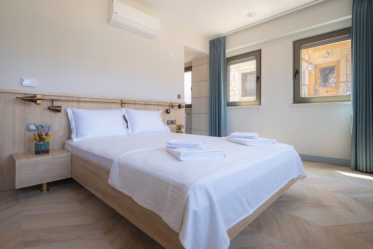 A modern hotel room with a large bed, wooden headboard, bedside tables, and a window with curtains.