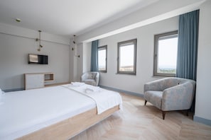 A modern hotel room with a large bed, a seating area with a chair, a TV mounted on the wall, and large windows offering a view.
