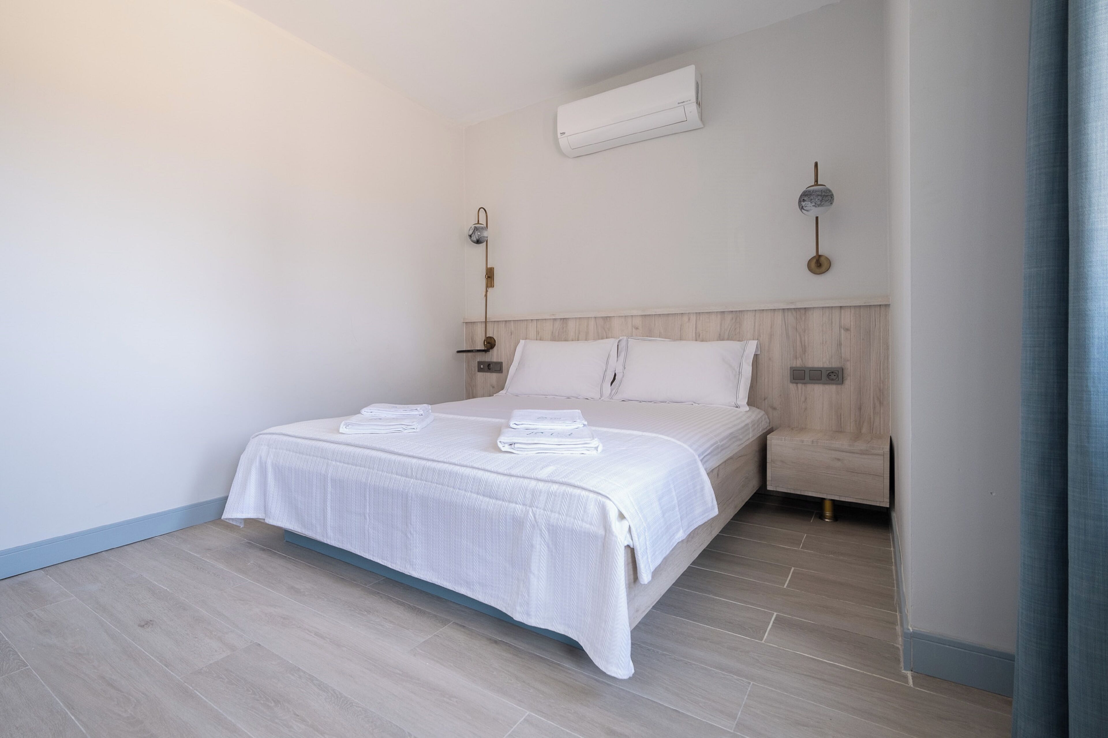 A neatly made bed with a white bedspread and pillows, a wooden headboard, a nightstand with a lamp, and an air conditioning unit on the wall.
