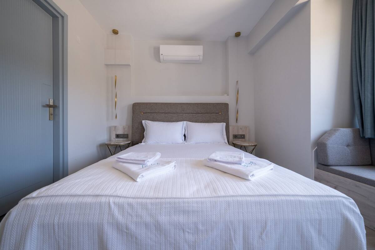 A neatly made bed with white linens and towels, a grey headboard, and bedside tables with lamps.