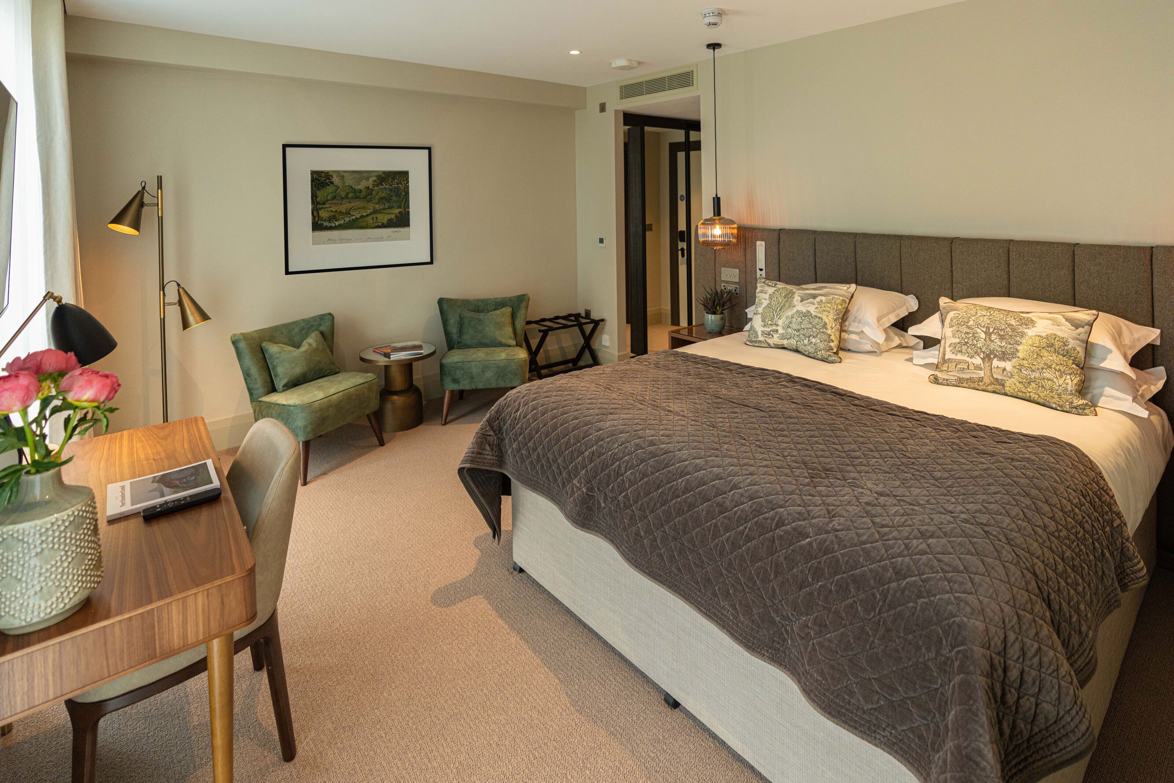 A hotel room with a large bed, a desk with a laptop, a chair, and a small table with a vase of flowers.