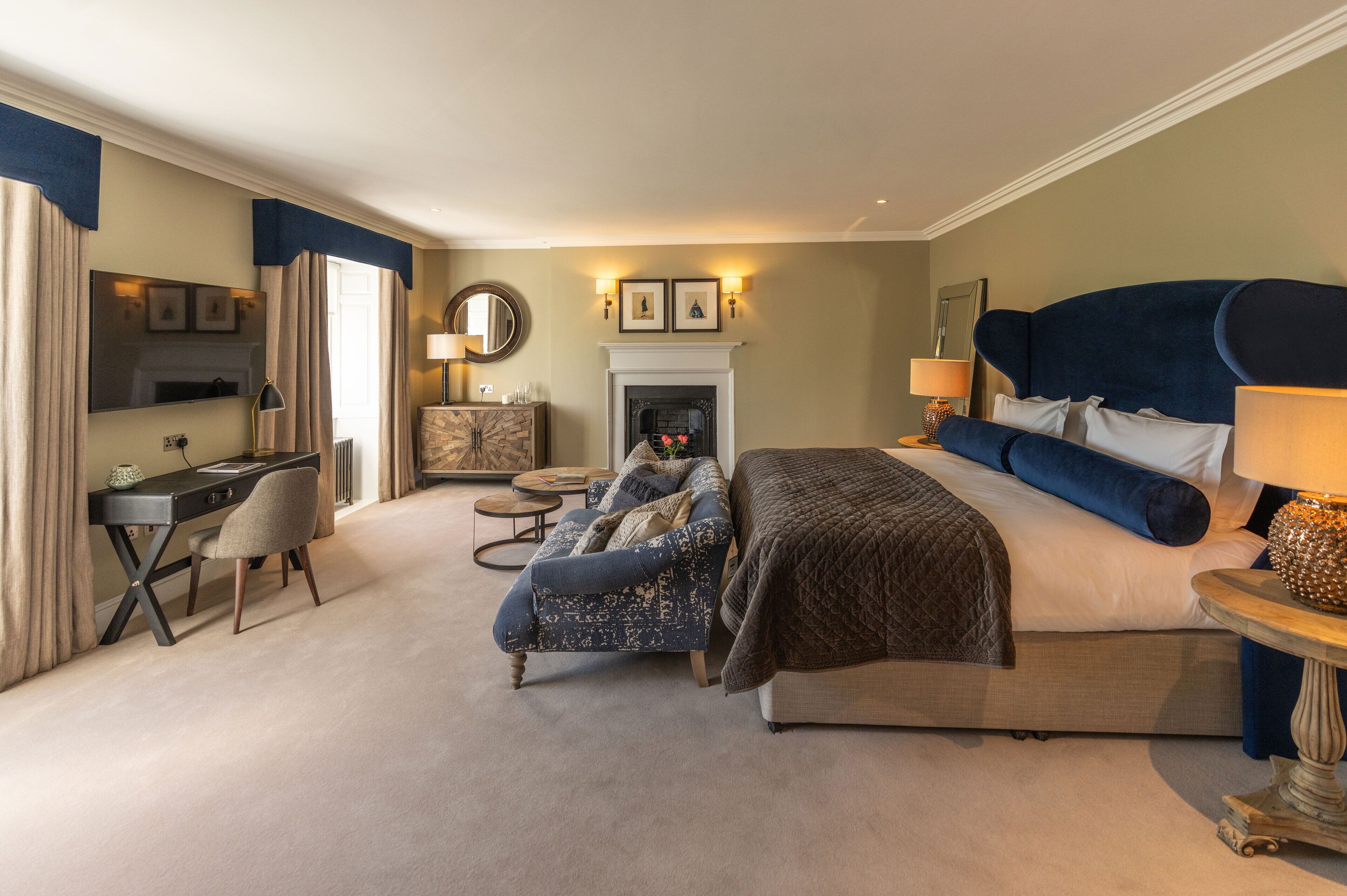 A hotel room with a large bed, a desk, a chair, a fireplace, and a mirror.