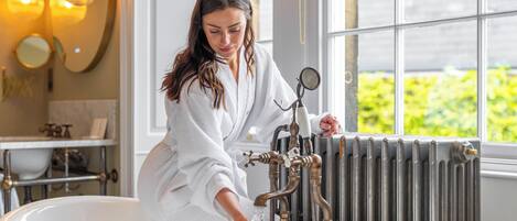 A person in a white bathrobe at a vintage-style bathtub with a radiator behind it, in a room with a large window and a mirror on the wall.