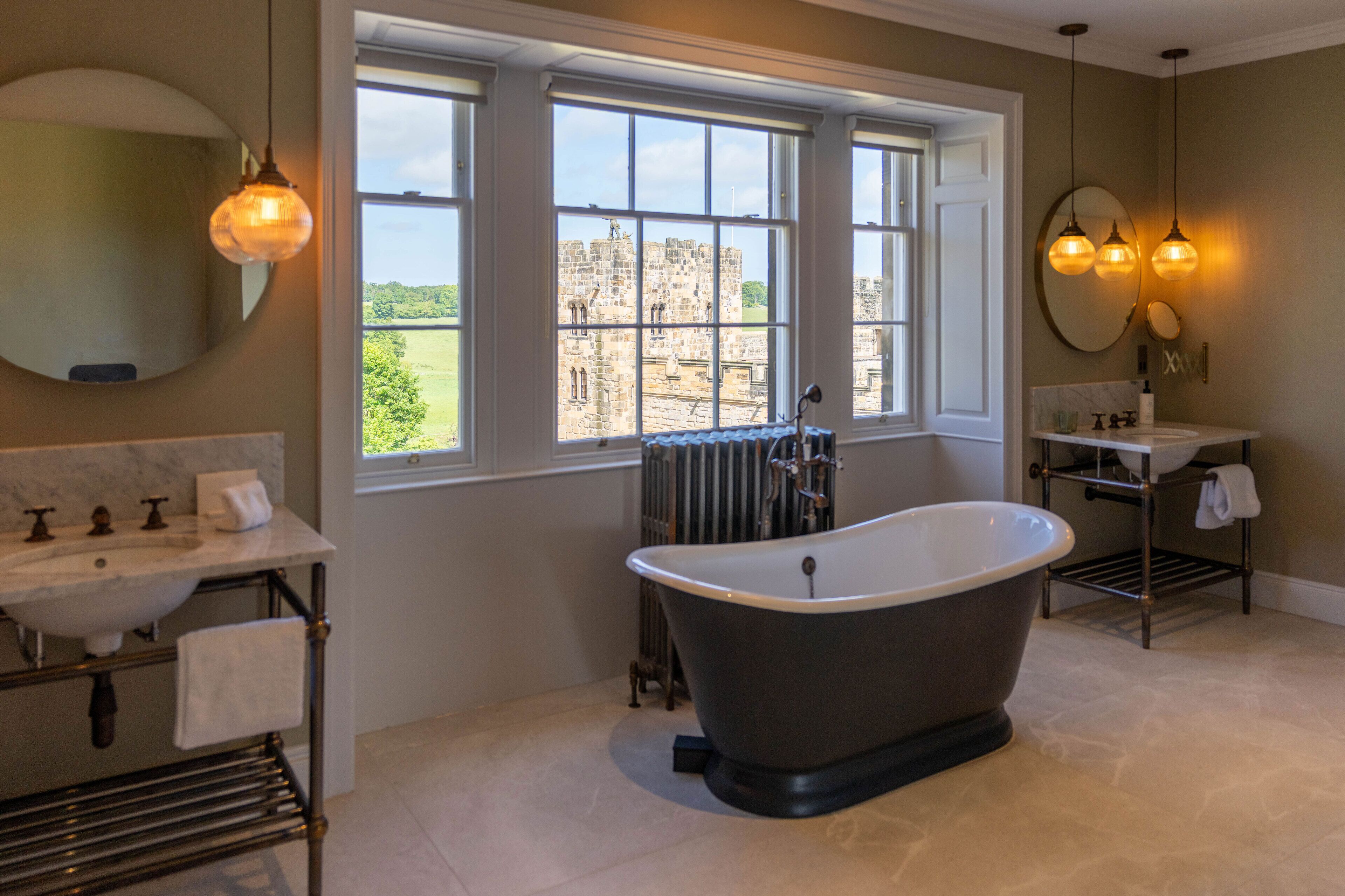 A bathroom with a freestanding bathtub, a sink with a marble countertop, a window with a view of a castle, and pendant lights.