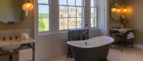 A bathroom with a freestanding bathtub, a sink with a marble countertop, a window with a view of a castle, and pendant lights.