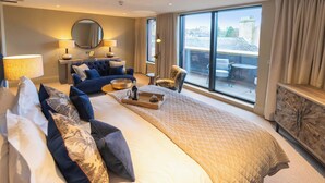 Habitación de hotel con una cama grande, una zona de estar con sofá y sillones, una mesa de comedor con bandeja, y vistas de la ciudad a través de amplios ventanales.