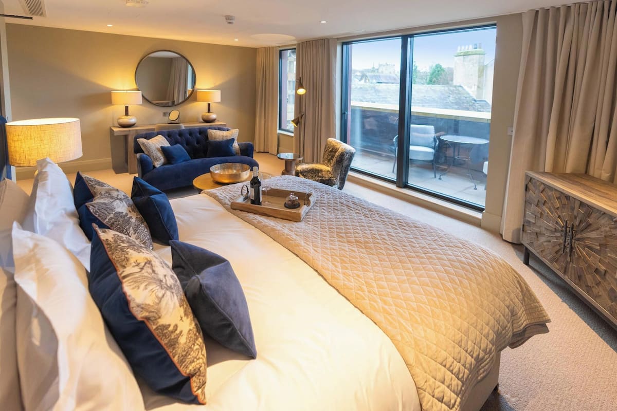 Une chambre d’hôtel dotée d’un grand lit, d’un coin salon avec un canapé et des fauteuils, d’une table à manger avec un plateau, et offrant une vue sur la ville grâce à de grandes fenêtres.