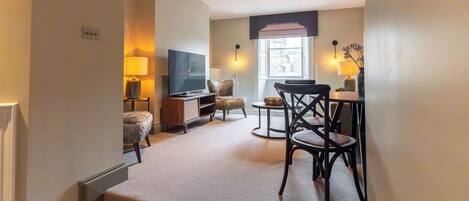 A hotel room with a flat-screen TV, a sofa, a dining table with chairs, a small rug, and a window with a view.