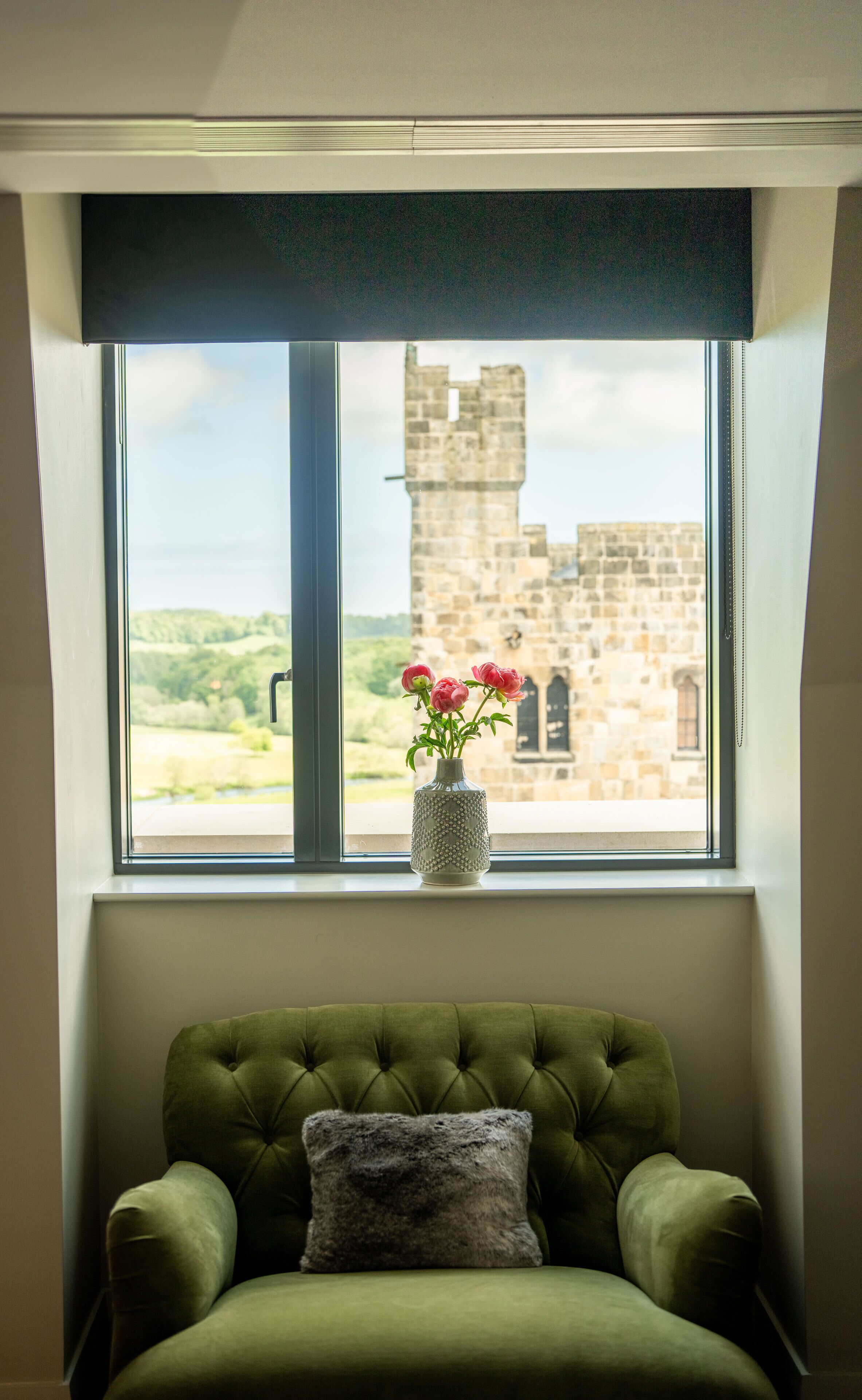 Una poltrona verde rivestita, con un cuscino decorativo, un vaso con fiori e vista su un edificio in pietra attraverso una finestra con tenda scura.