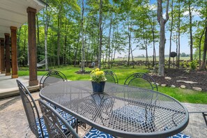 A covered patio with a round metal table and chairs, a potted plant, and a view of a forested area. - Modern Egg Harbor Home | Dog-Friendly + Fire Pit (Egg Harbor)