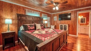 A bedroom with a wooden ceiling, a bed with a quilt, a nightstand, a lamp, a TV, and a kitchen area visible through an open door.