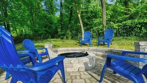A circular stone fire pit with four blue Adirondack chairs arranged around it in an outdoor setting.