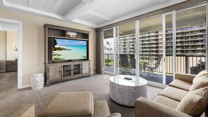 Un salón amplio con un televisor de pantalla plana grande, asientos cómodos y un balcón con vistas al océano y un edificio de gran altura.