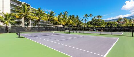 Cancha de tenis con red, rodeada de palmeras y un edificio.
