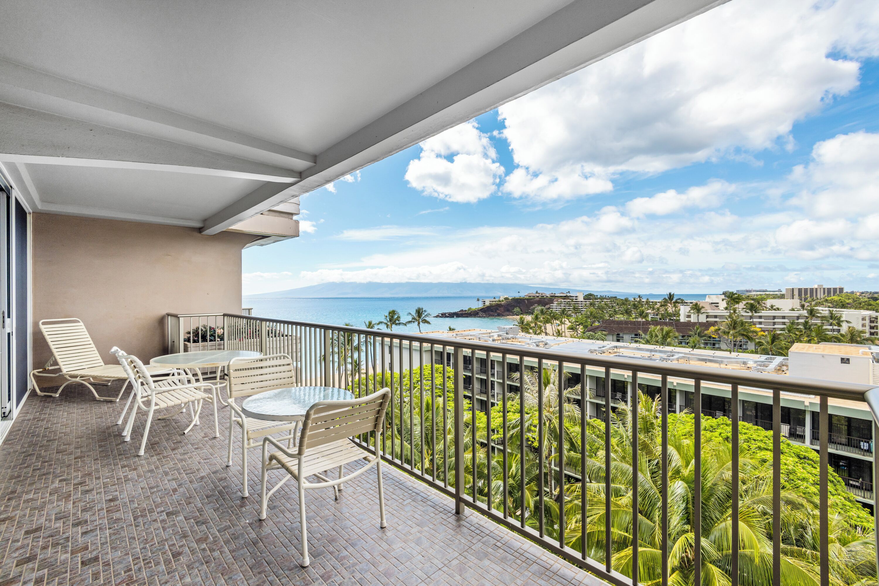 A balcony with a table and chairs, overlooking a coastal view with palm trees and buildings.