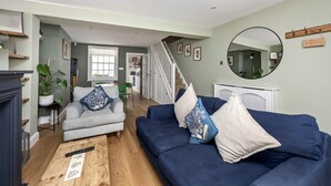 Ein geräumiges Wohnzimmer mit einem blauen Sofa, einem grauen Sessel und einem Holztisch. Eine Treppe führt zu einer oberen Etage, und ein runder Spiegel befindet sich an der Wand.