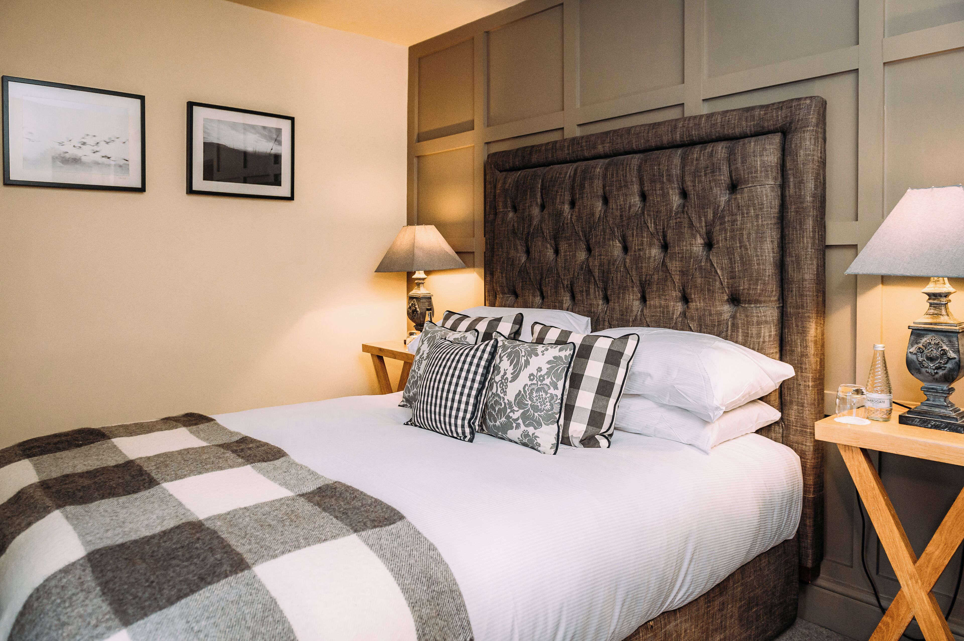 A neatly made bed with a checkered blanket, a tufted headboard, and multiple pillows. There are two framed pictures on the wall, a bedside lamp, and a small table with a bottle and glass.