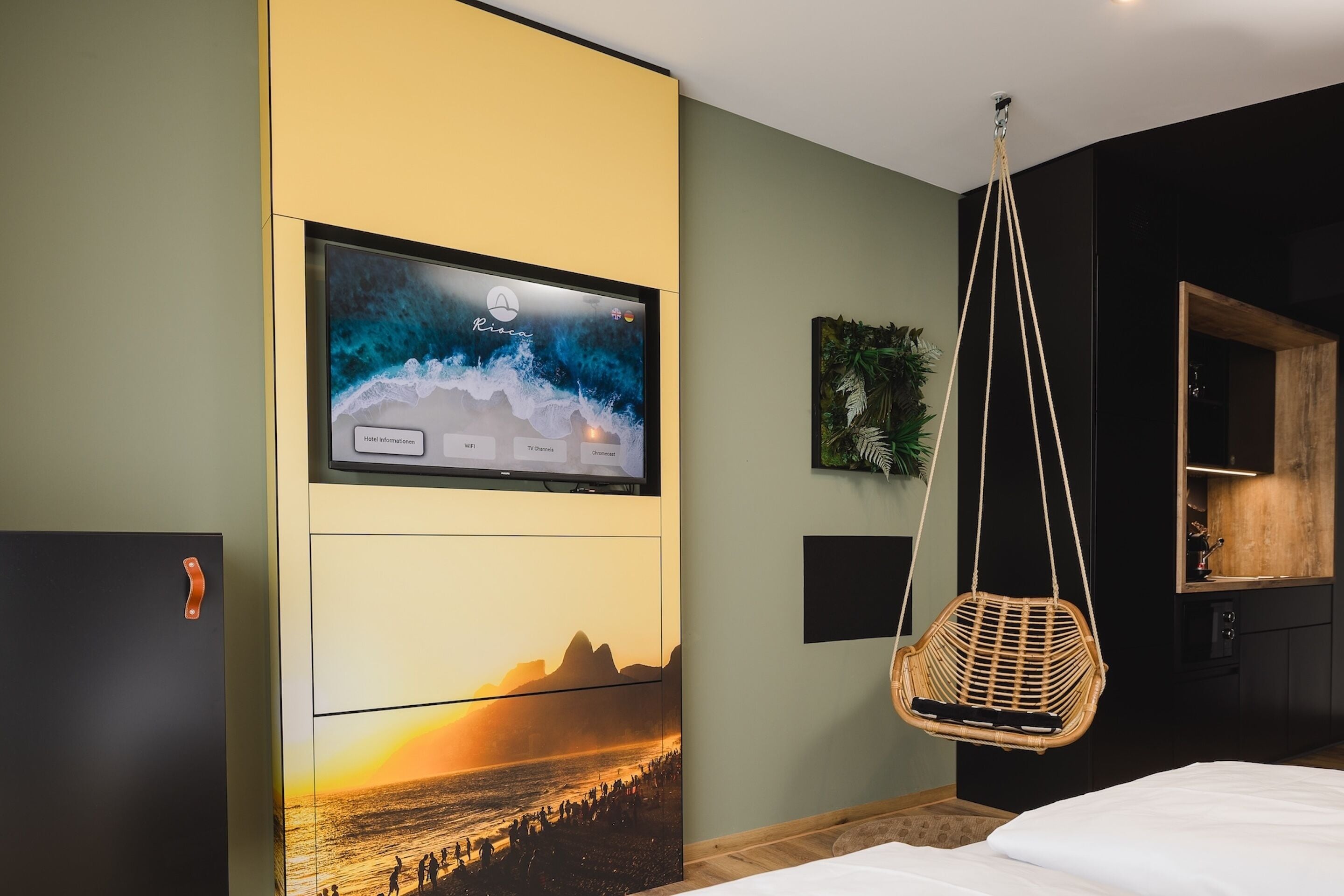 Une chambre d’hôtel moderne dotée d’un grand téléviseur fixé au mur qui diffuse une image panoramique, d’un fauteuil suspendu en rotin et d’un lit recouvert de draps blancs.