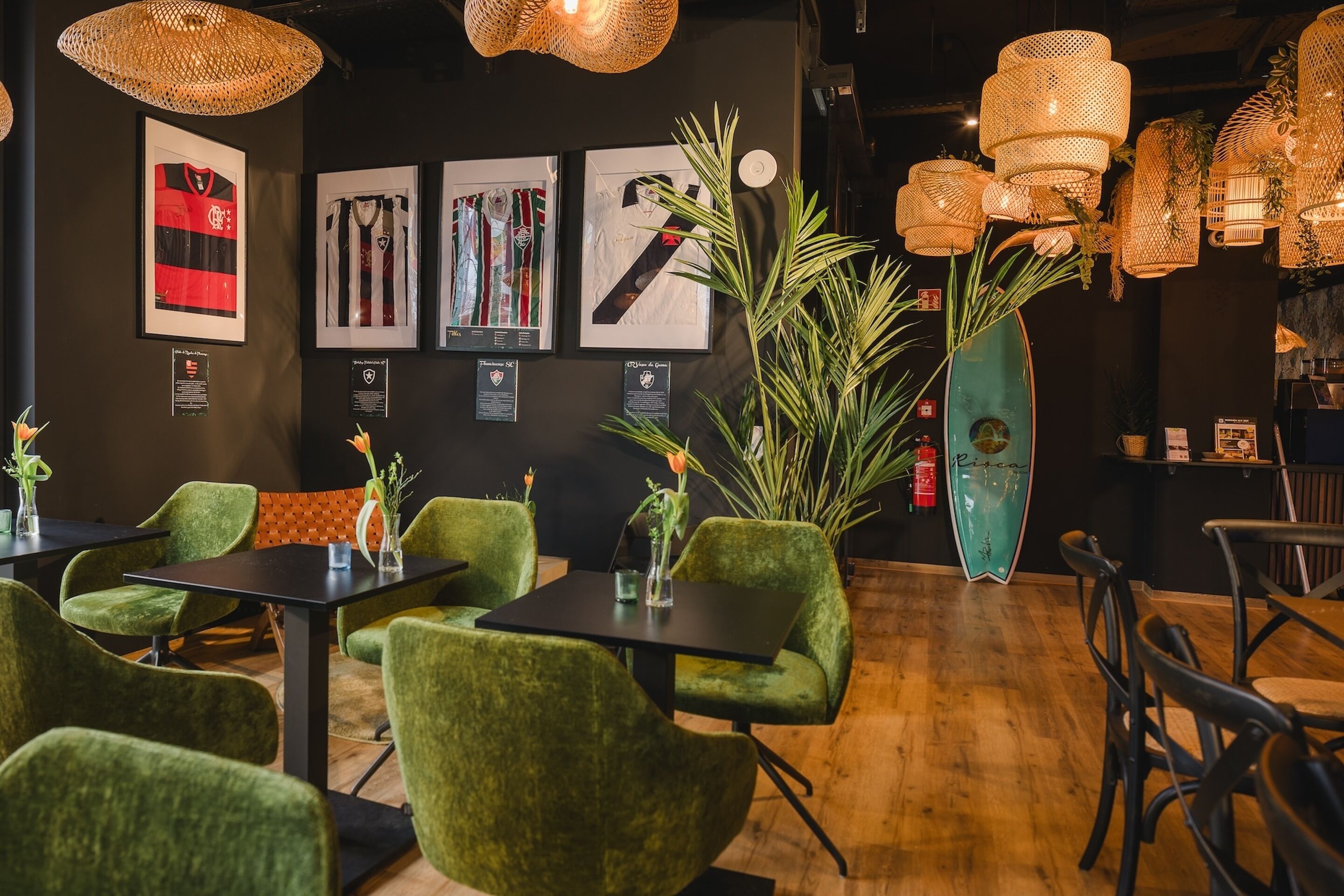 Interior de restaurante con sillas tapizadas de verde, mesas negras y camisetas deportivas enmarcadas en la pared.