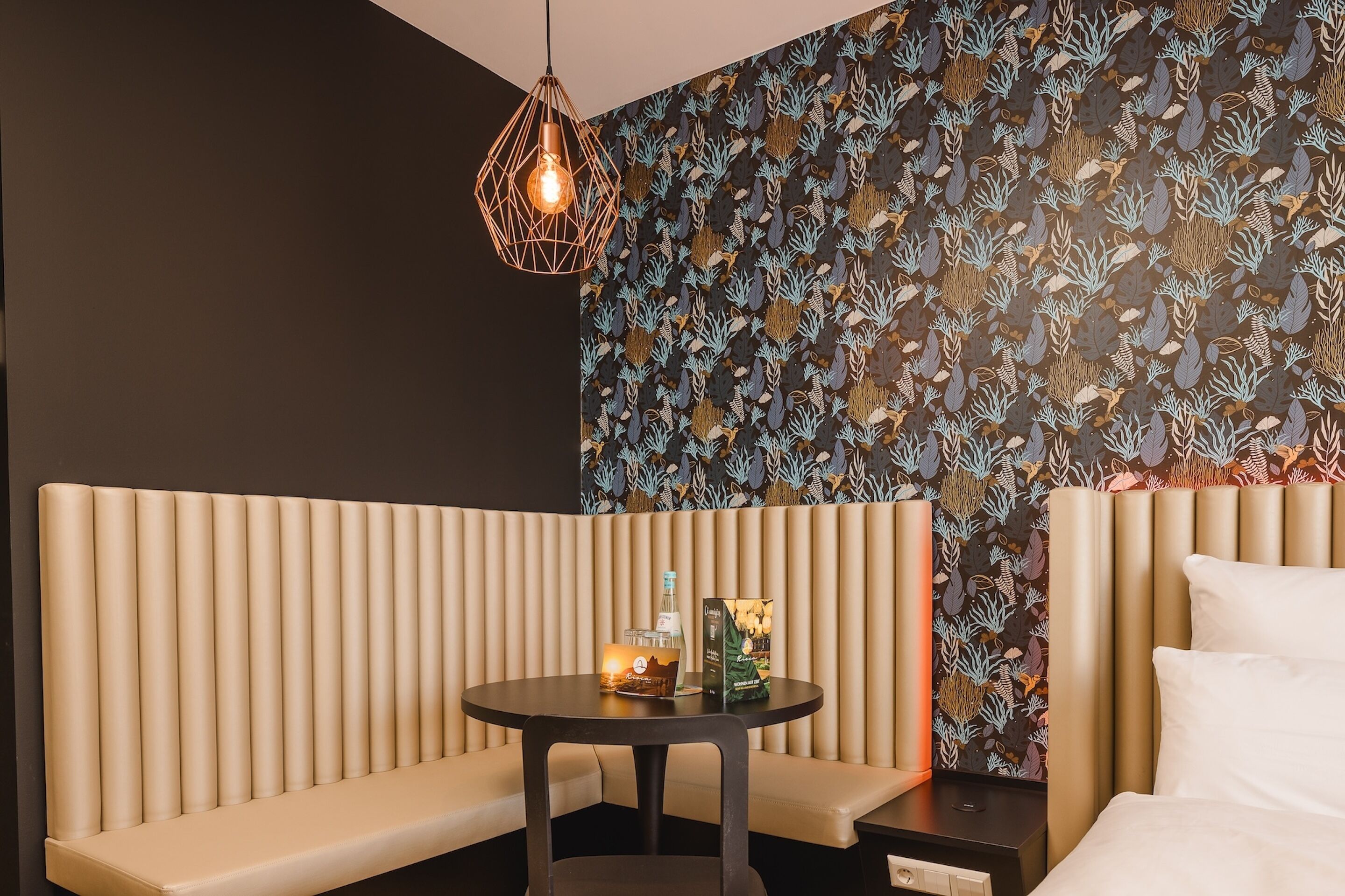 Une chambre d’hôtel moderne avec un banc rembourré, une petite table ronde sur laquelle se trouvent une bouteille et une boîte, et un papier peint à motifs.