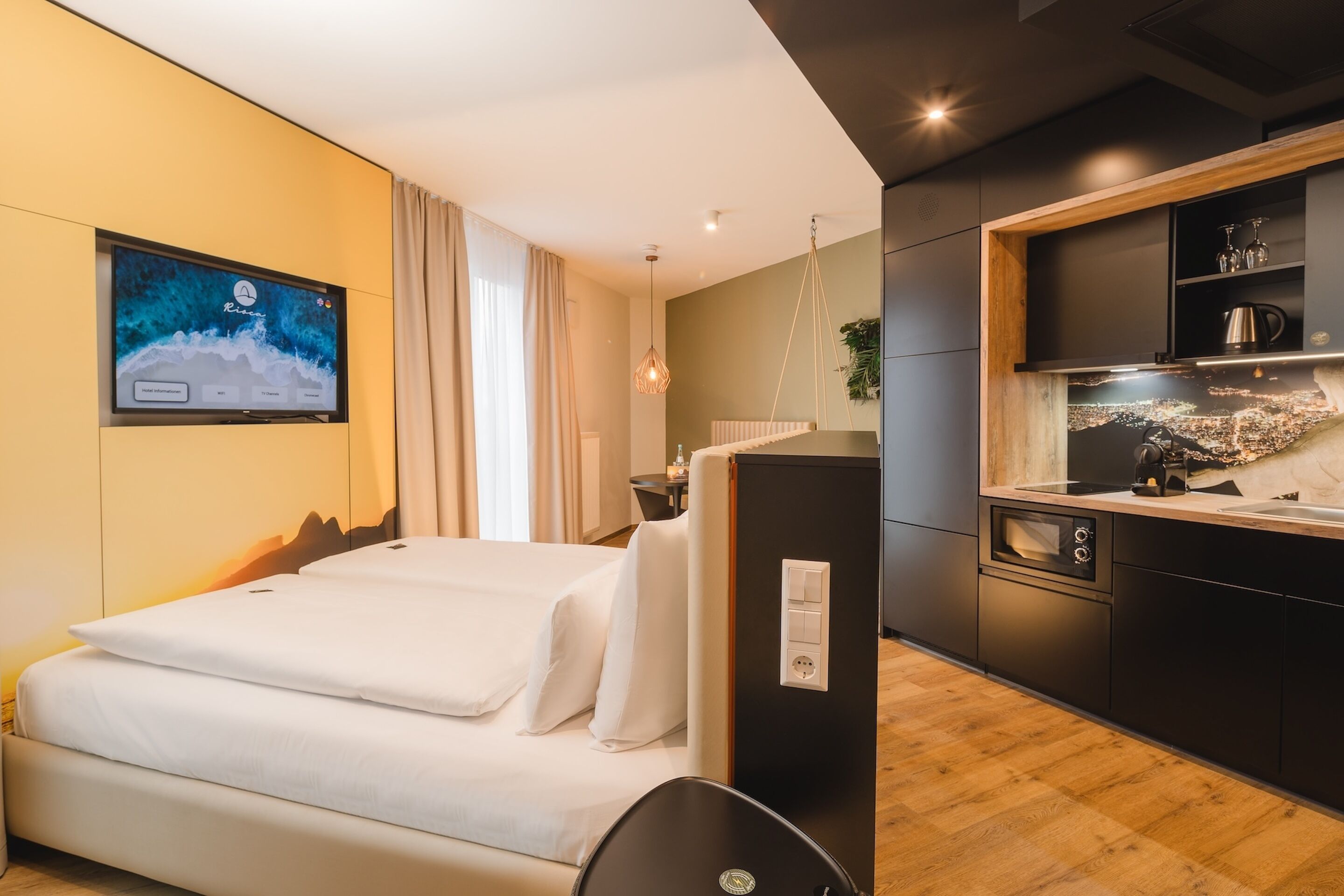 Una habitación de hotel moderna con una cama, un televisor montado en la pared, una pequeña zona de cocina con un fregadero y armarios, y suelo de madera.