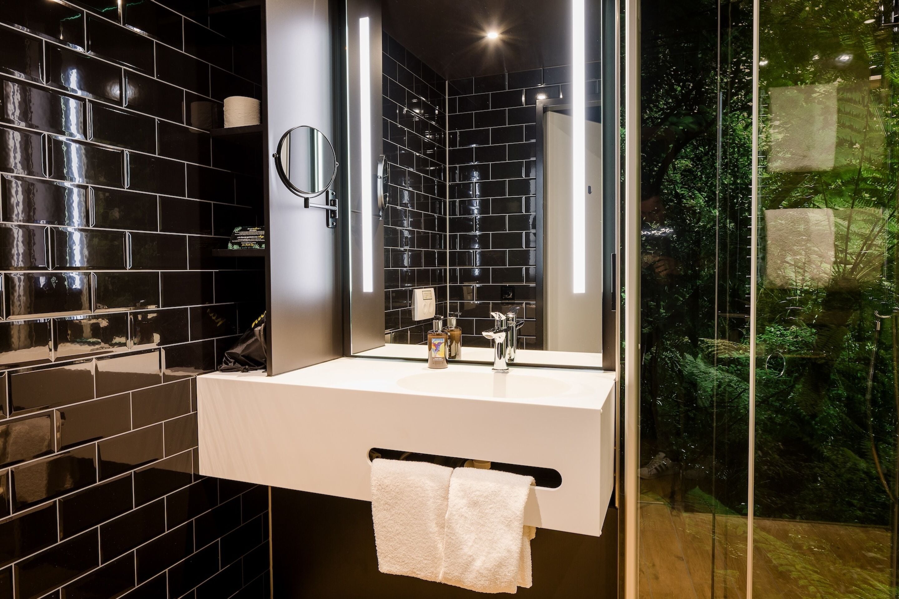 A modern bathroom with black tiled walls, a white sink, a mirror, and a glass shower enclosure with a view of greenery.