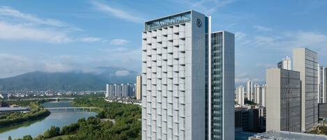 A modern hotel with a prominent tower and a shorter building, surrounded by a river and greenery.