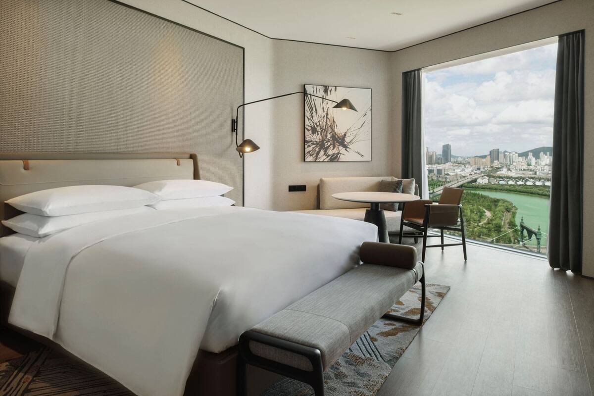 Une chambre d’hôtel moderne dotée d’un grand lit, d’un coin salon avec une table et offrant une vue sur le paysage urbain grâce à de grandes fenêtres.