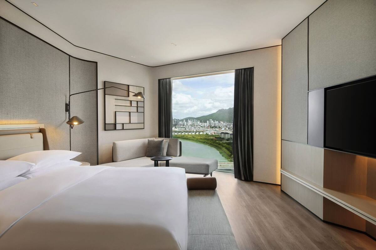 Une chambre d’hôtel moderne dotée d’un grand lit, d’un canapé, d’une télévision et offrant une vue sur le paysage urbain grâce à une grande fenêtre.