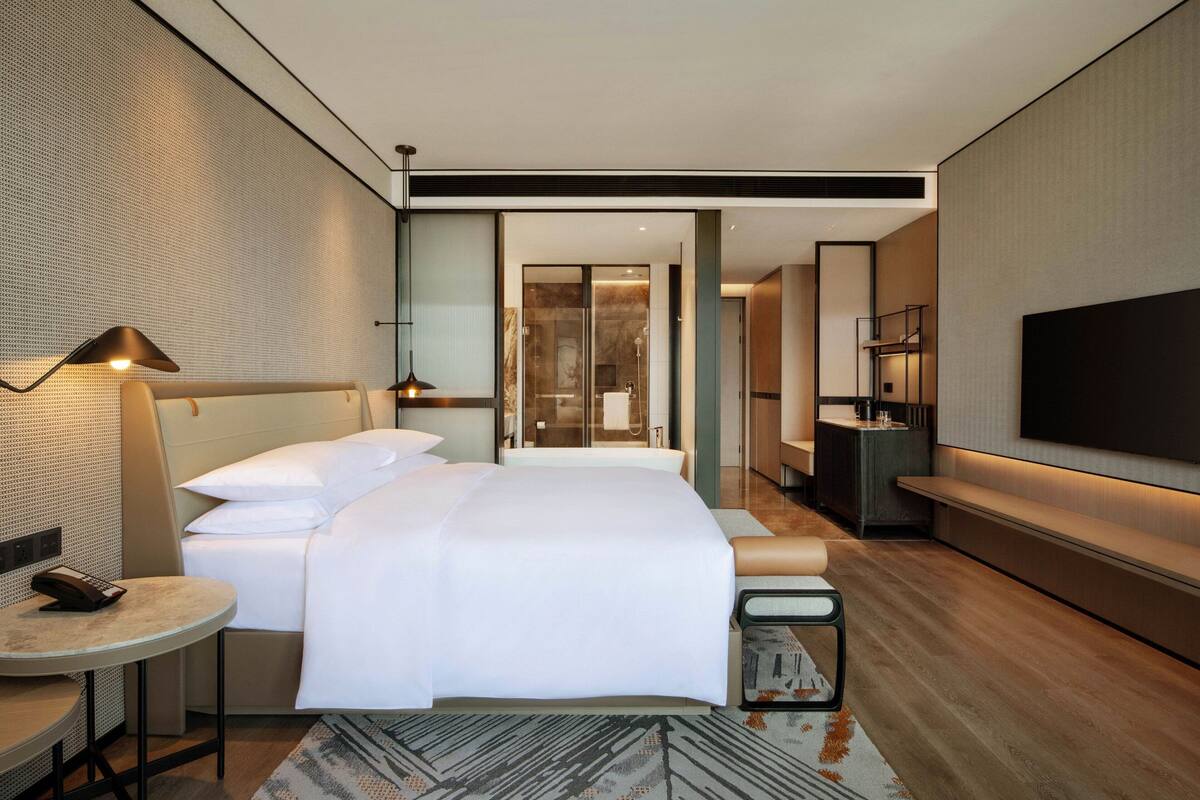 Une chambre d’hôtel moderne avec un grand lit, des tables de chevet, une télévision et une salle de bain visible à travers une porte en verre.