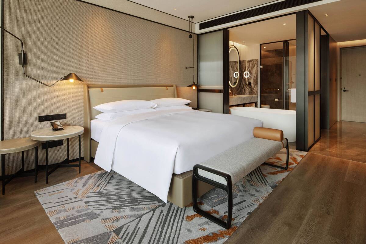 Une chambre d’hôtel moderne avec un grand lit, une table de chevet avec une lampe, une petite table ronde et une salle de bain visible à travers une porte en verre.