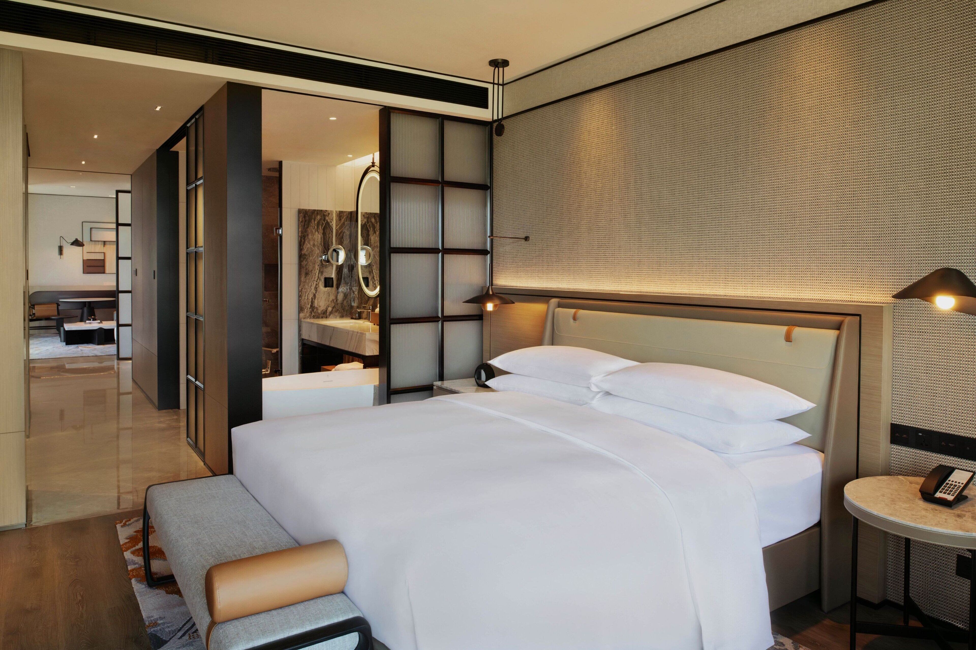 A modern hotel room with a large bed, a bedside table with a lamp, a mirror, and a view of the hallway.