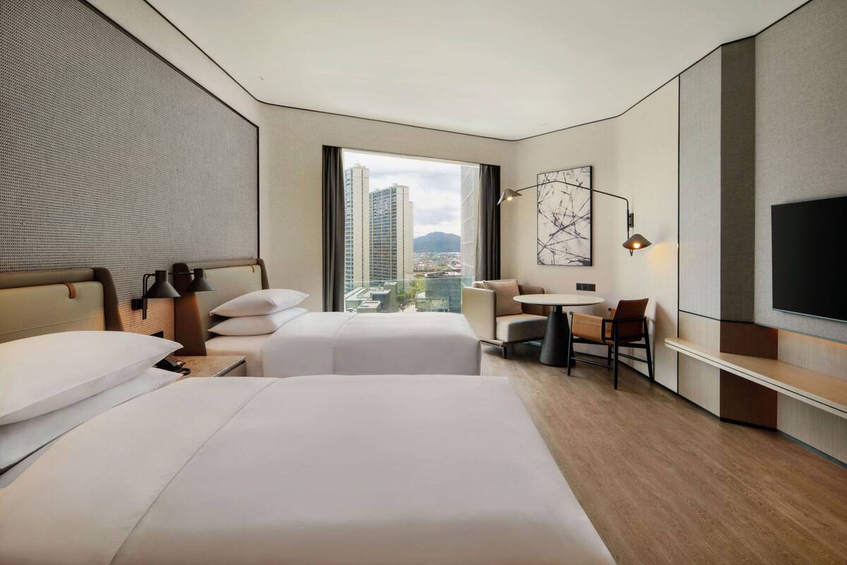 Une chambre d’hôtel moderne équipée de deux lits, d’un bureau, d’une chaise, d’une télévision et d’une grande fenêtre donnant sur la ville.