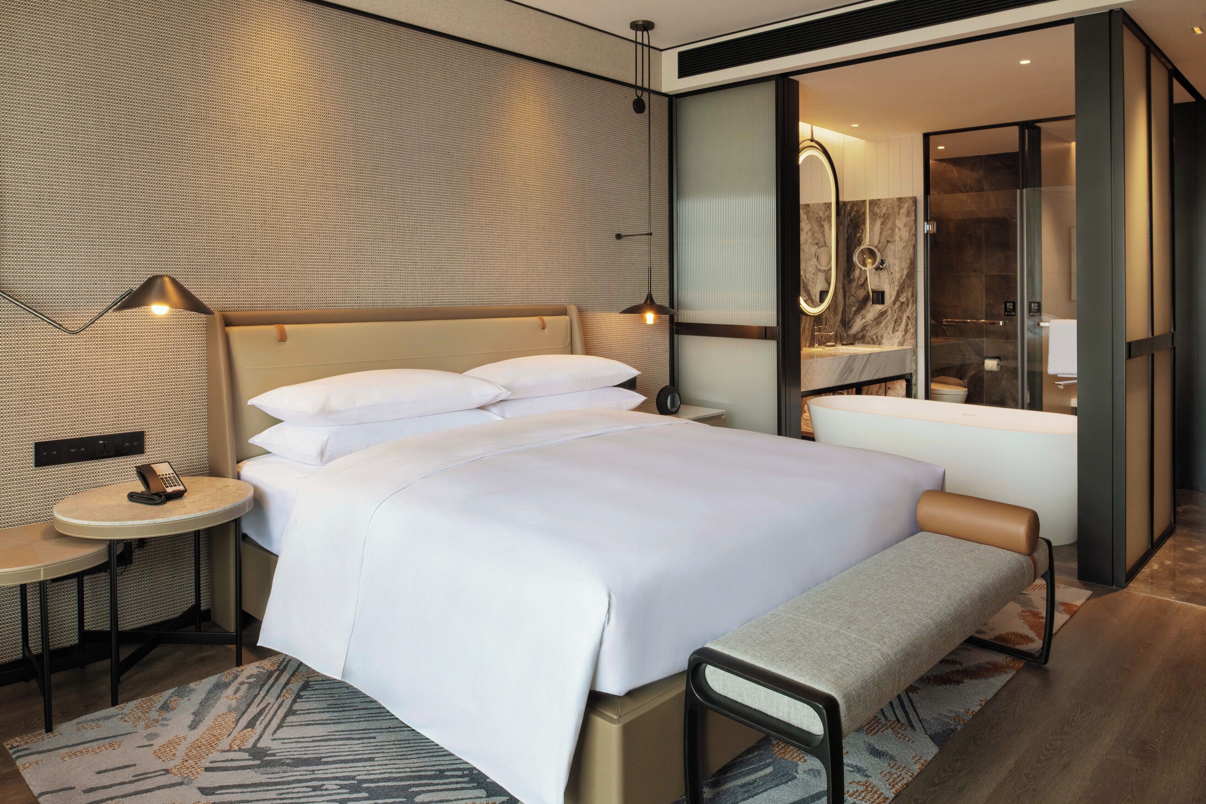 A modern hotel room with a large bed, bedside tables, a bench, a shower area, and a small table with a phone.