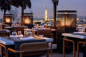 Un restaurante en la azotea con mesas dispuestas para comer, con vistas a un paisaje urbano donde destaca una torre.