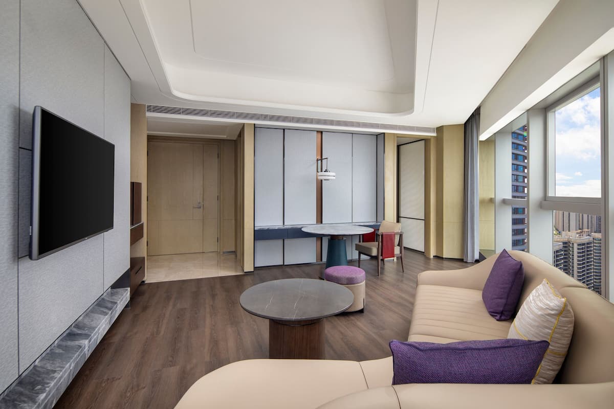 A modern living room with a flat-screen TV, a round coffee table, a sofa with cushions, a dining area with a table and chairs, and a view of high-rise buildings through large windows.