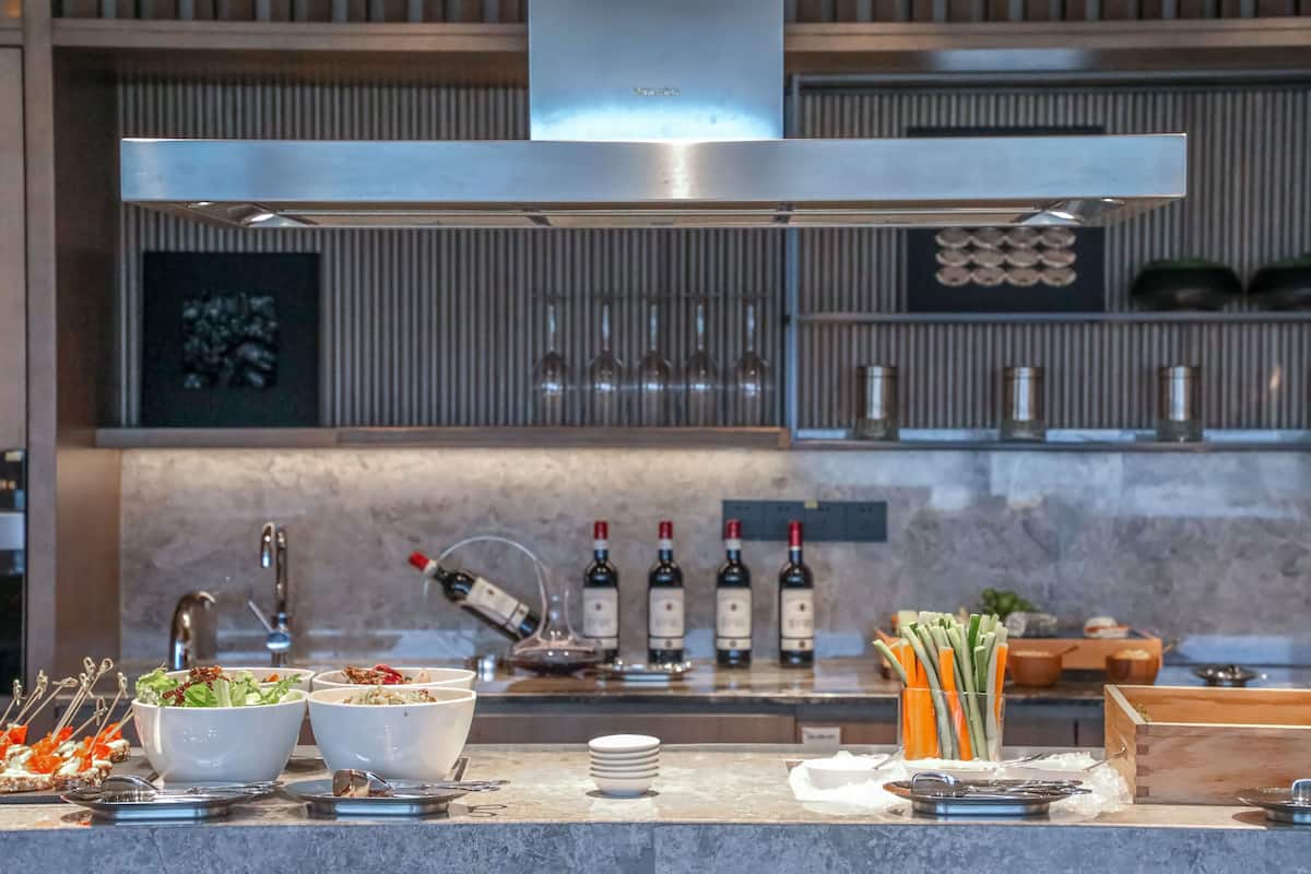 A modern kitchen with a stainless steel range hood, a wine rack with bottles, and various food items on the counter.