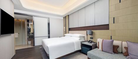 A modern hotel room with a large bed, a sofa, a flat-screen TV, and a walk-in closet.