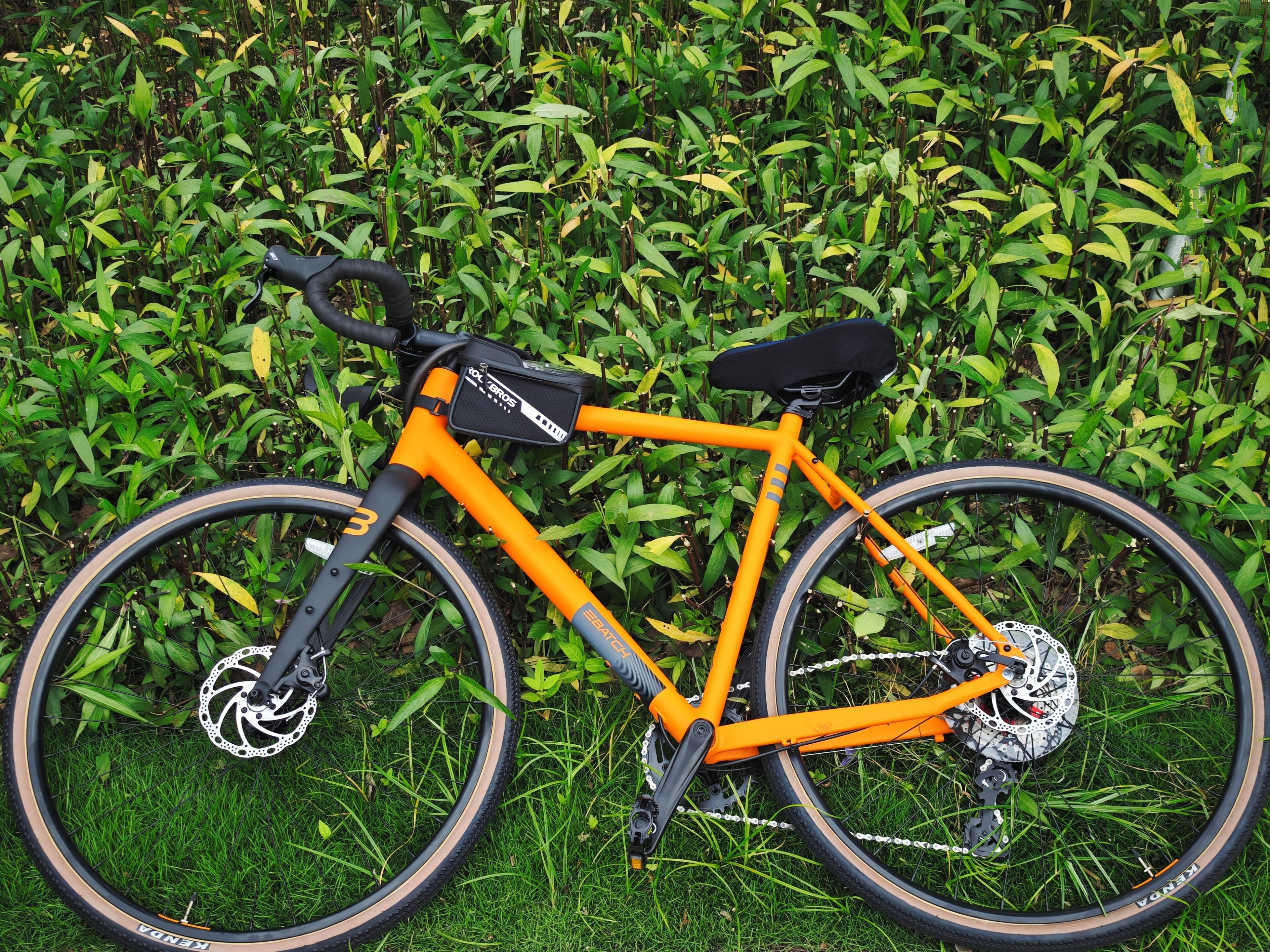 Een oranje fiets, stilstaand, met een zwart zadel en een waterfleshouder op het frame, geparkeerd op gras met een weelderige groene haag op de achtergrond.