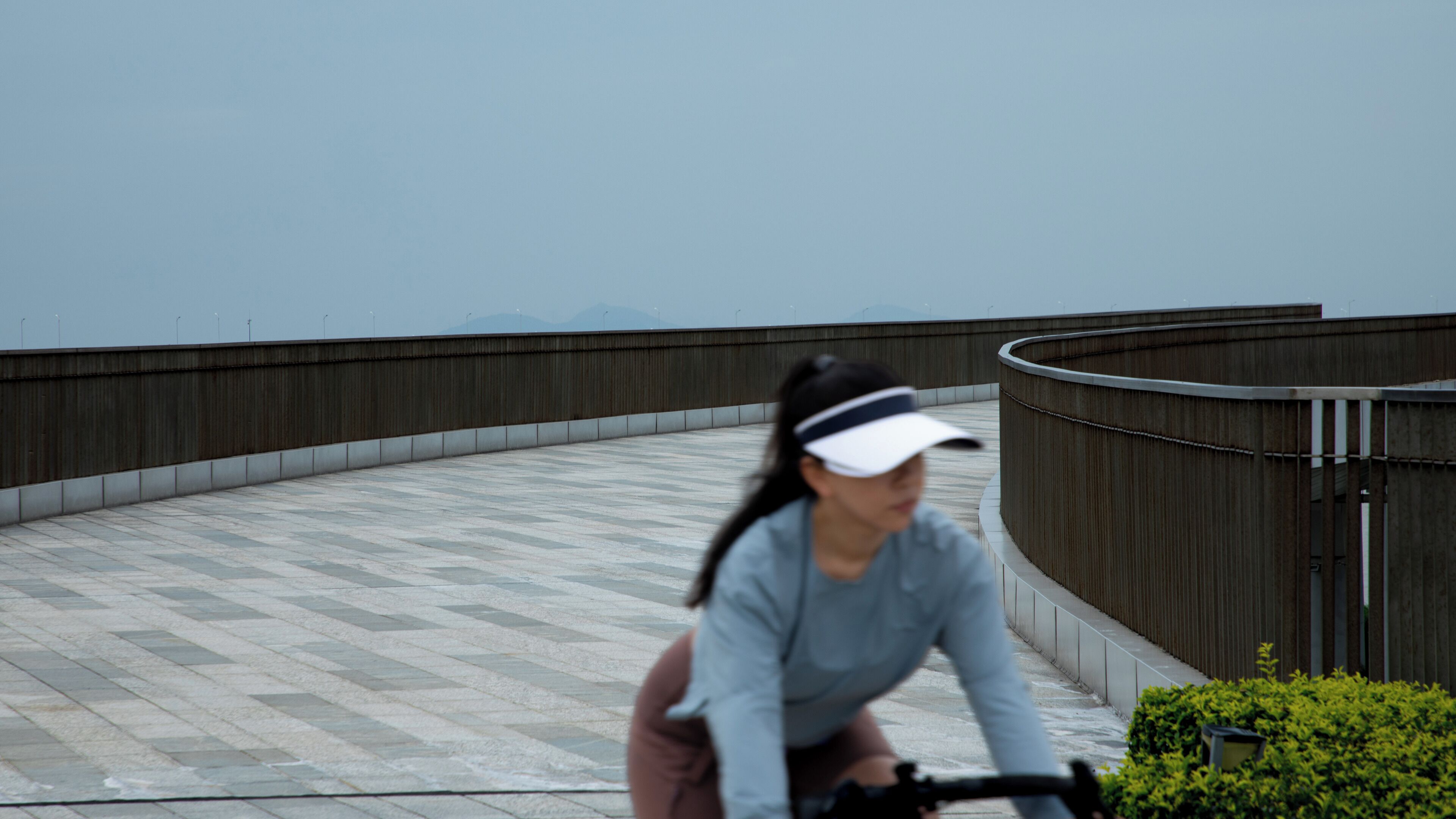 A person riding a bicycle on a curved pathway with a concrete surface, surrounded by a low wall and a metal fence.
