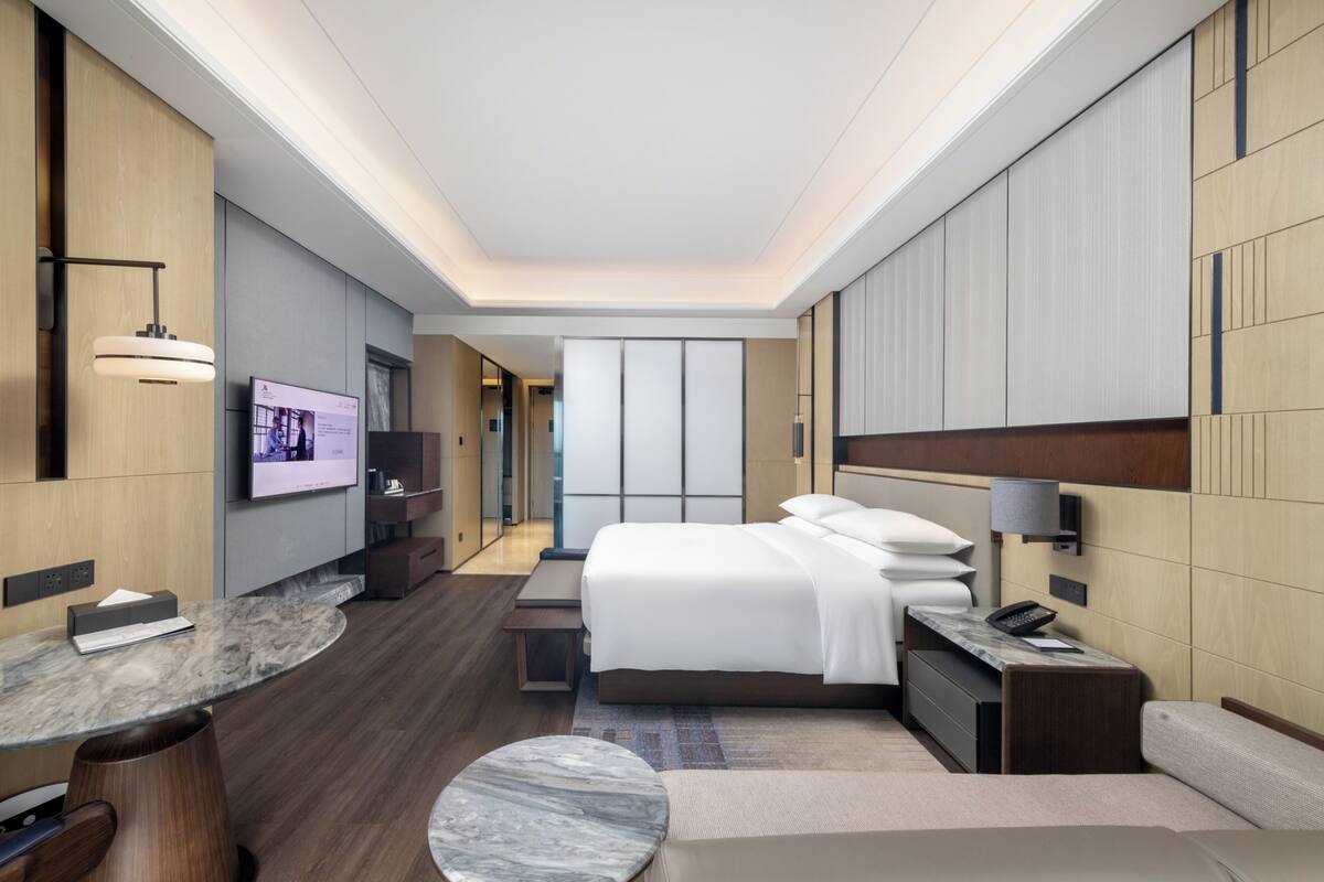 A modern hotel room with a large bed, a desk, a TV, and a seating area.