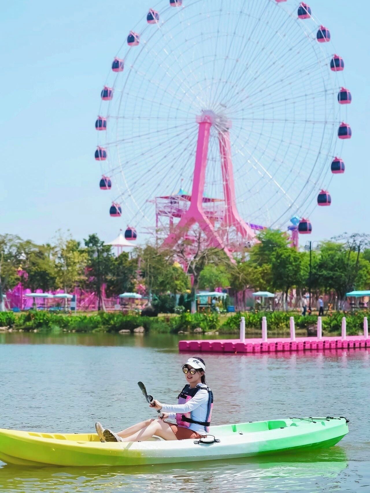 A person in a green kayak on a body of water with a large pink Ferris wheel in the background.