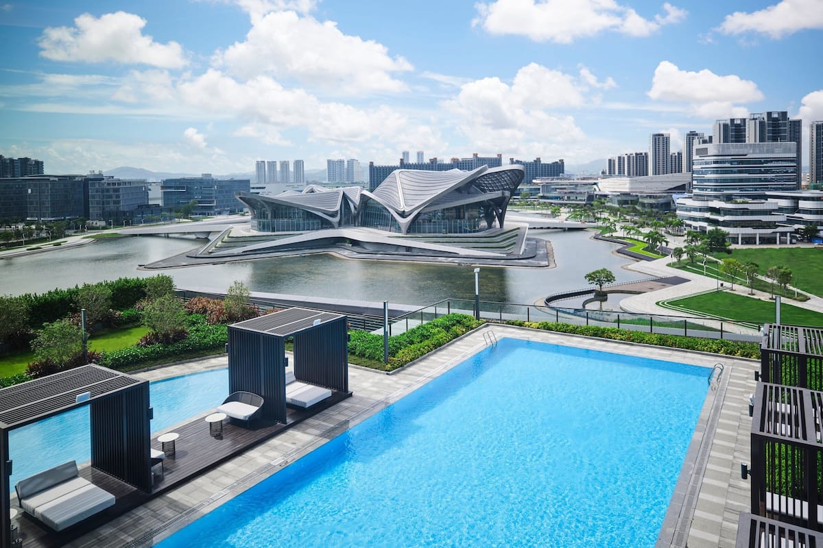 A modern hotel with a swimming pool, a river, and a distinctive building in the background.