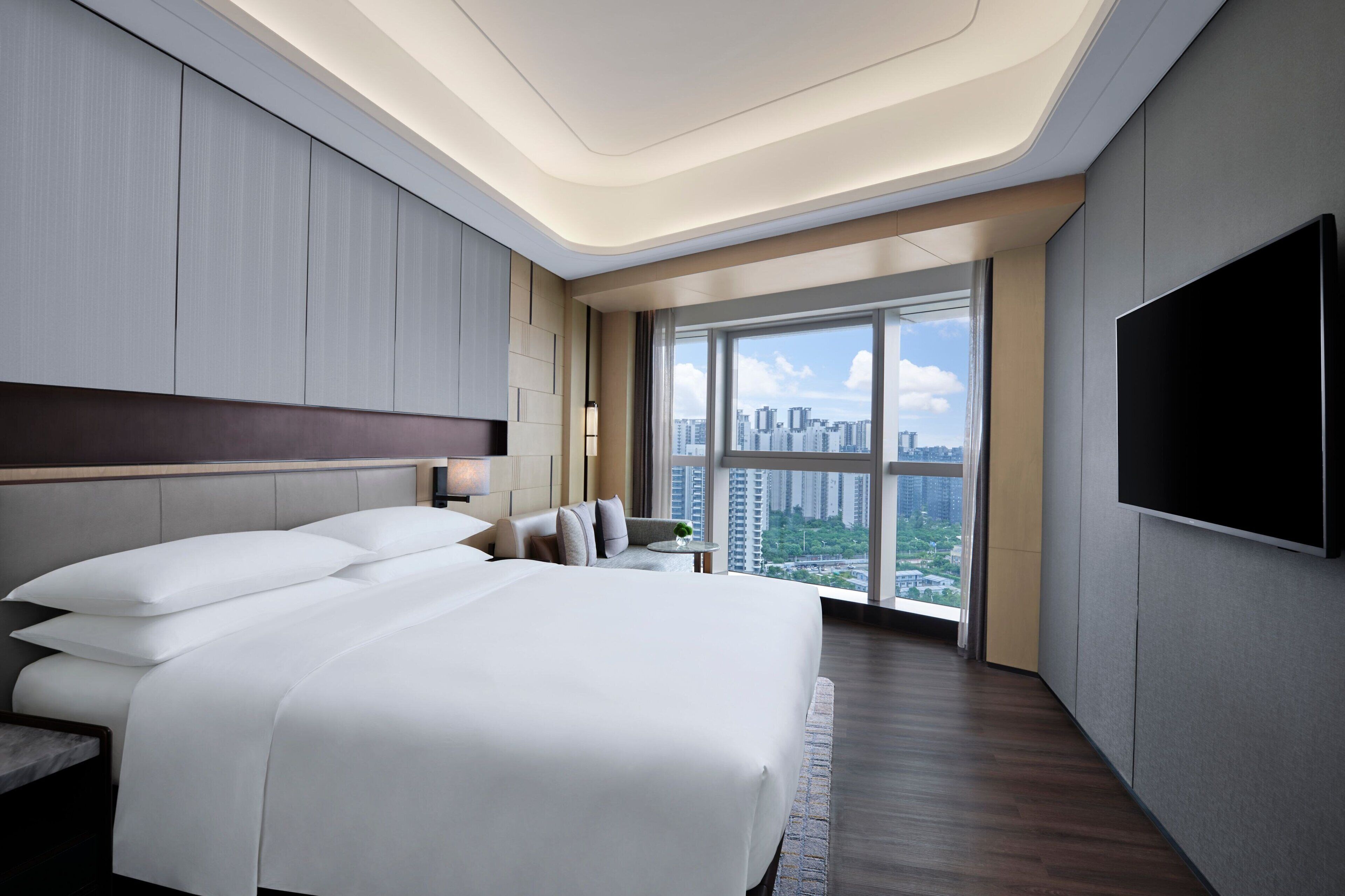 A modern hotel room with a large bed, a flat-screen TV, and a city view through large windows.