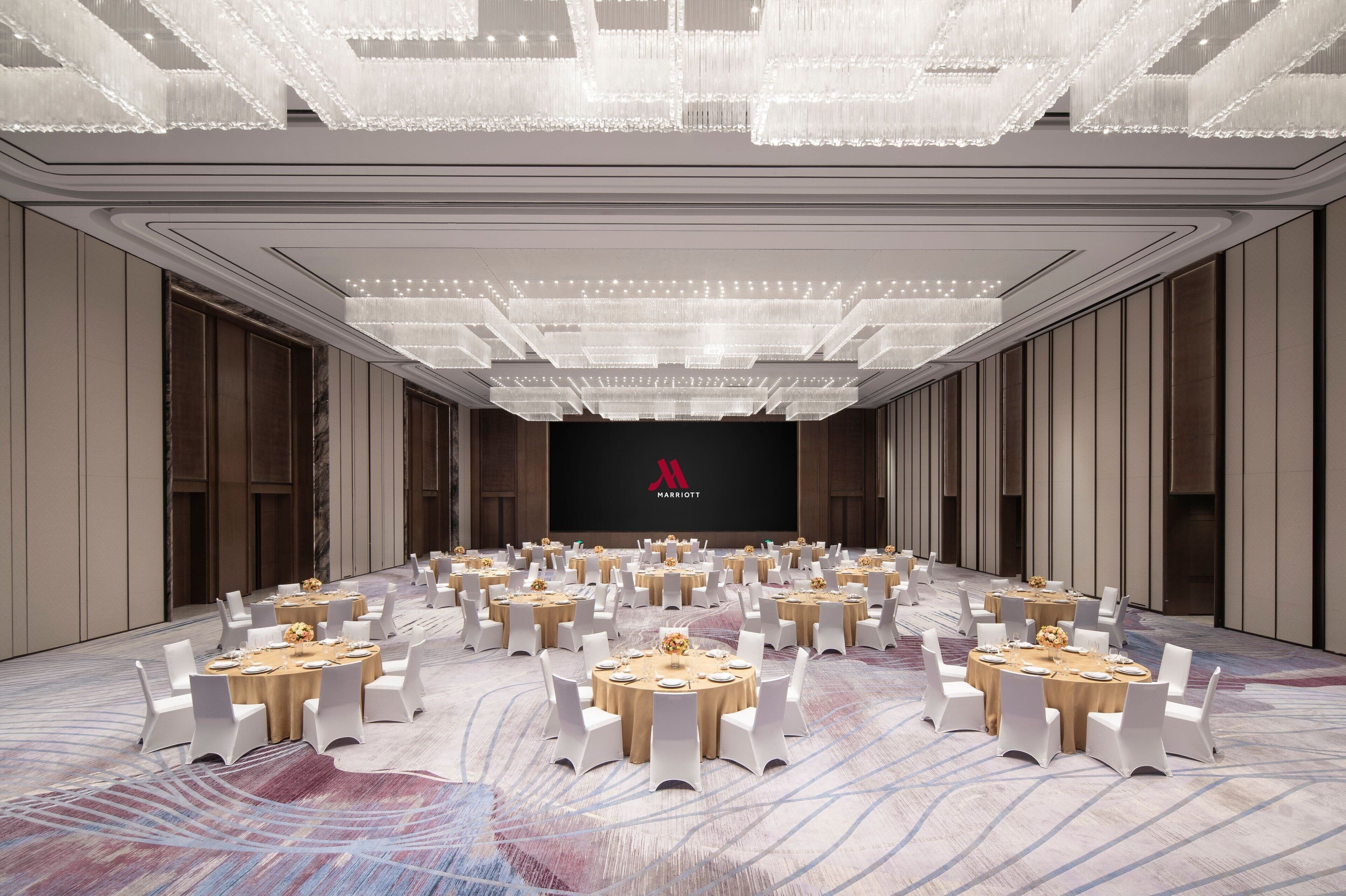 A spacious conference room with round tables and white chairs, a large screen with a logo, and a patterned carpet.
