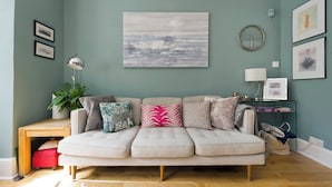 A living room with a light-colored sofa, a small wooden desk, a potted plant, and a wall-mounted lamp. - I Came To Boogie (London)