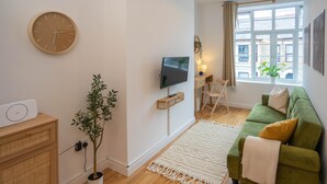 En moderne stue med en grøn sofa, et fladskærms-tv, et træskab, en potteplante og et stort vindue med udsigt til bygninger.