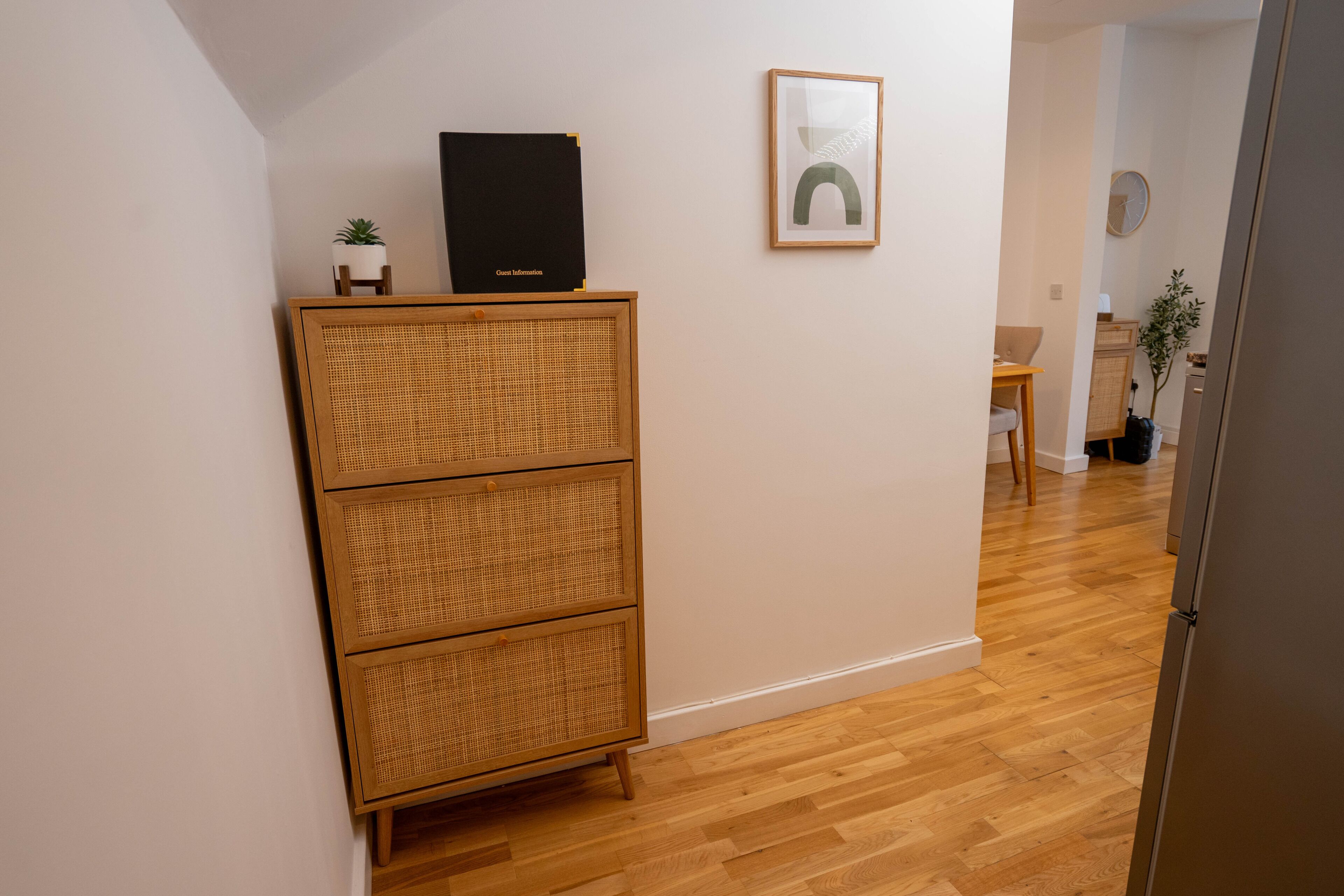 A hallway with a wicker cabinet, a framed artwork, and a door leading to another room.