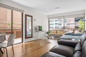 Ein modernes Wohnzimmer mit Sofa, Couchtisch und Sesseln, einem Fernseher und großen Fenstern mit Blick auf Gebäude.